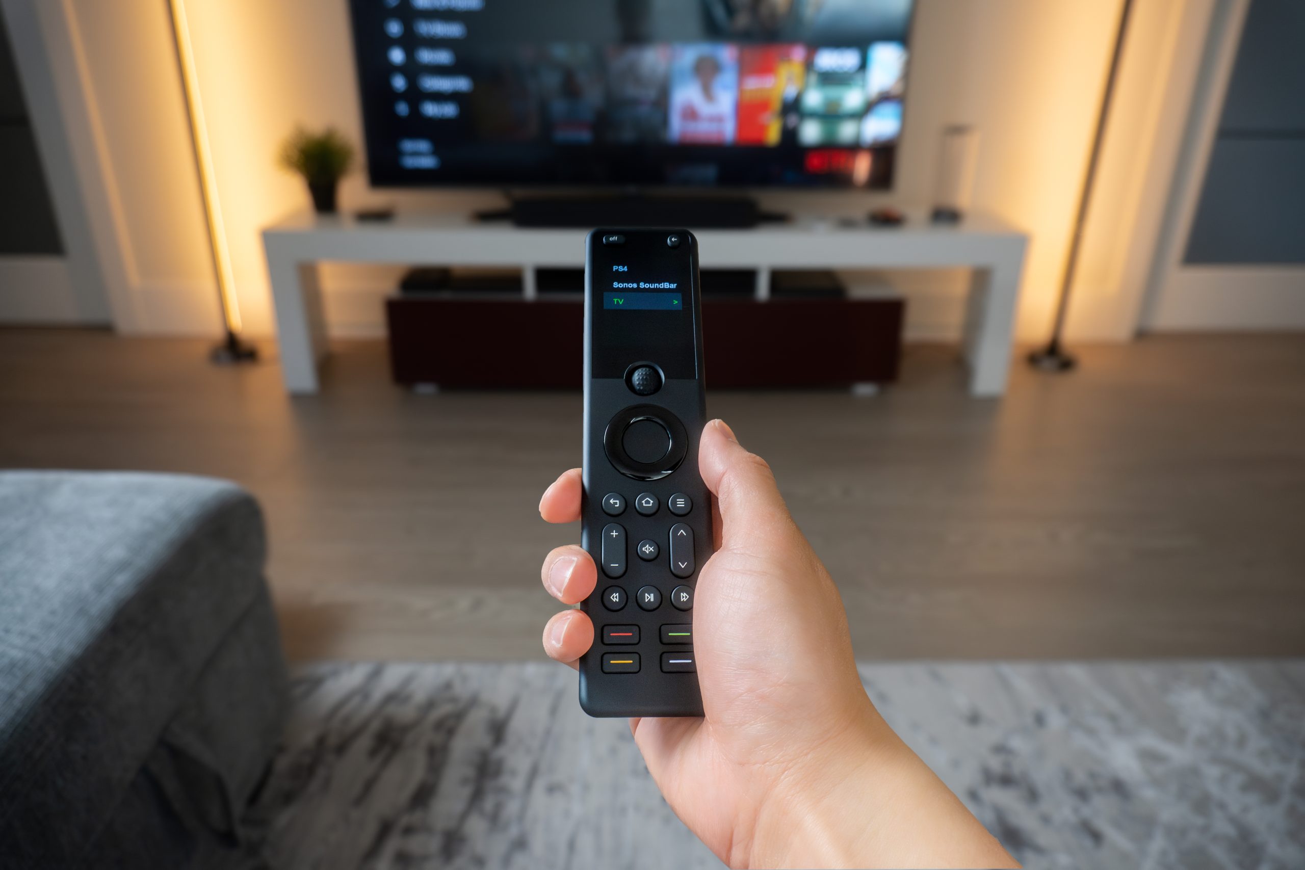 Hand using a SofaBaton X1S remote to select a TV, Sonos soundbar, or PS4.